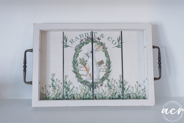 A white wooden tray with black handles features a floral wreath, carrots, and the text RABBIT & CO in green. Part of the Flipping Smalls Easter Edition, it&rsquo;s adorned with delicate white flowers and greenery along the edges.