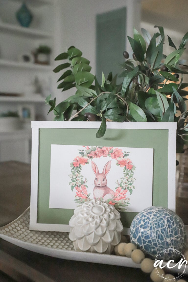 A framed illustration of a rabbit surrounded by flowers sits on a tray with a decorative white flower, a blue patterned ball, and green leaves in this cozy, softly lit room&mdash;perfect for an Easter Edition of Flipping Smalls.