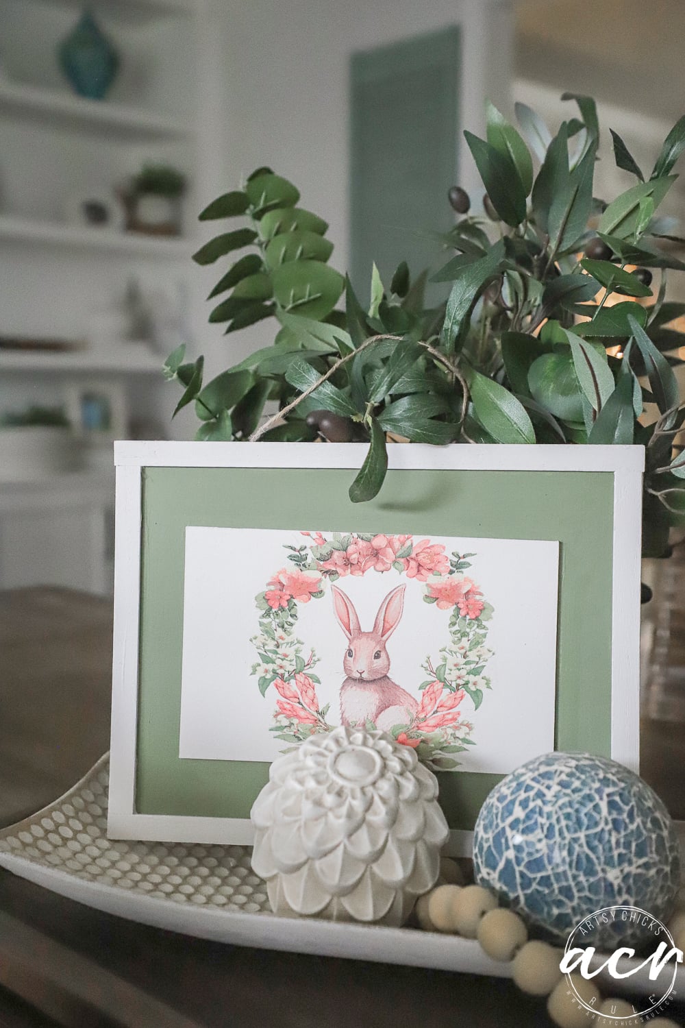 A framed illustration of a rabbit surrounded by flowers sits on a tray with a decorative white flower, a blue patterned ball, and green leaves in this cozy, softly lit room&mdash;perfect for an Easter Edition of Flipping Smalls.