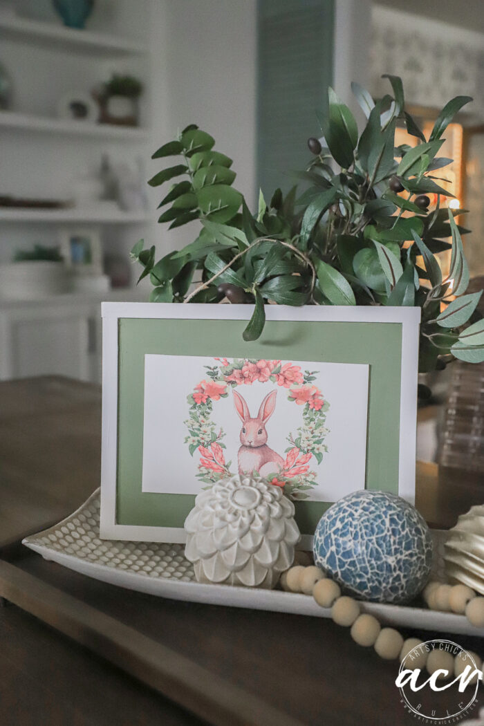 A decorative tray on a table holds a framed bunny illustration with a floral wreath&mdash;perfect for an Easter Edition&mdash;alongside a potted leafy plant, textured balls, wooden bead garland, and a white carved artichoke ornament.
