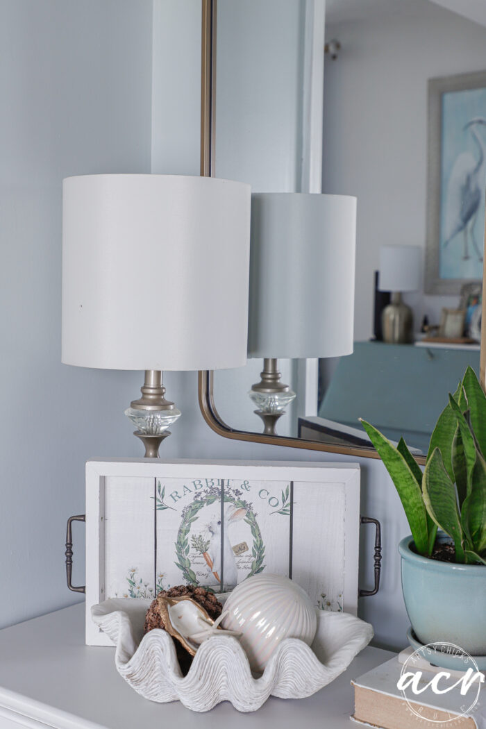 A white table holds a decorative clam shell with seashells, a potted plant, and a white lamp. Behind the shell rests a white tray with floral art&mdash;an Easter Edition touch&mdash;while the mirror reflects this Flipping Smalls coastal-themed room.