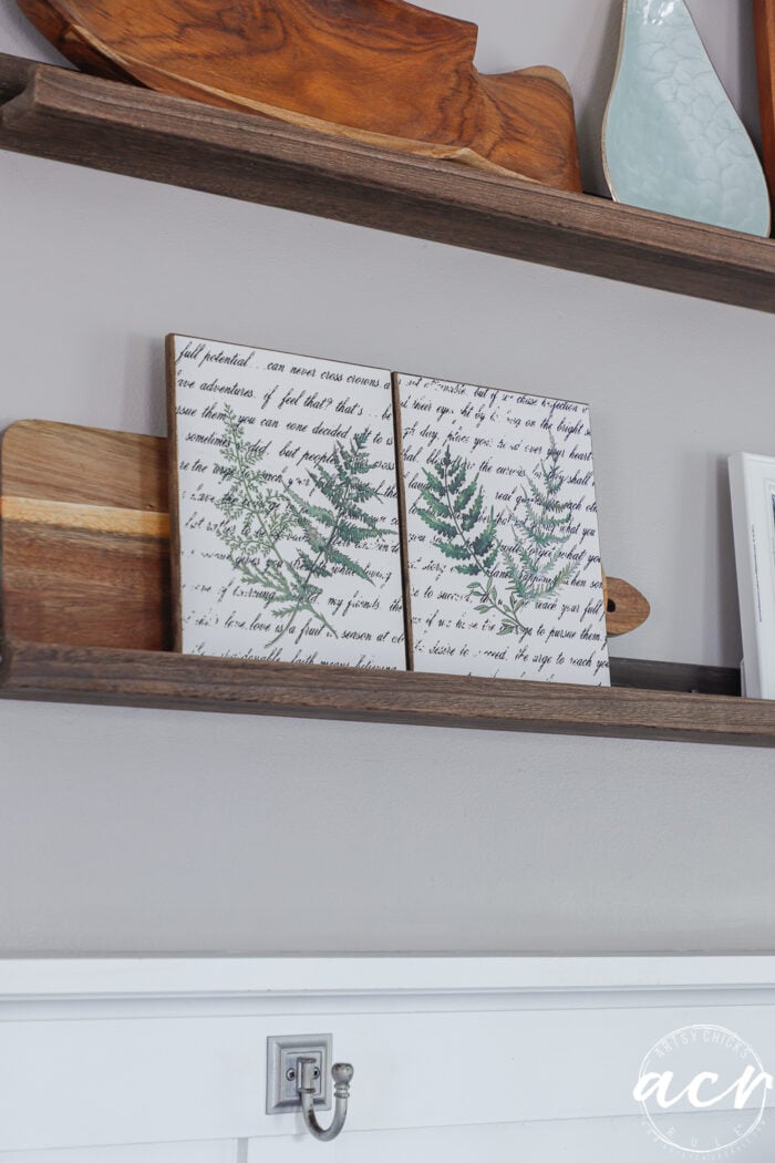 Two wooden shelves display decorative items, including a green leaf-printed artwork on script background, wooden cutting boards, and ceramic pieces&mdash;all arranged for this Easter Edition of Flipping Smalls against a gray wall with a silver hook below.