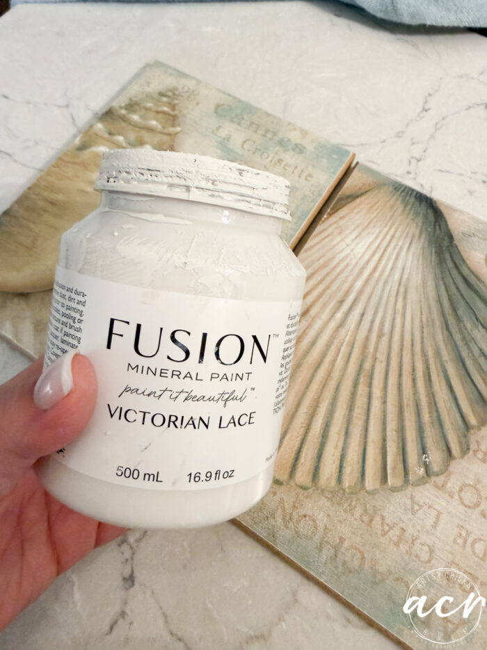 A hand holds a jar of Fusion Mineral Paint in the color Victorian Lace above a decorative surface featuring large seashell designs, perfect for an Easter Edition project or your next Flipping Smalls creation, set against a soft, neutral background.