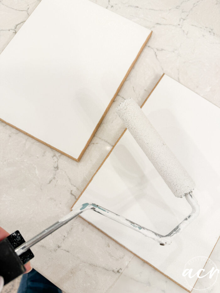 A hand holds a paint roller applying white paint to a blank canvas on a marble surface, with another blank canvas nearby&mdash;perfect for your next Flipping Smalls: Easter Edition creative project.