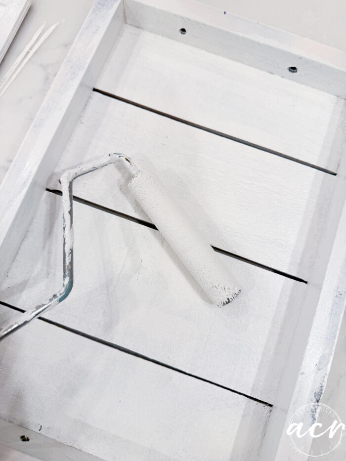 A small paint roller covered in white paint rests inside a white wooden tray with slats. The tray appears freshly painted, adding a clean, bright touch&mdash;perfect for a Flipping Smalls Easter Edition refresh.