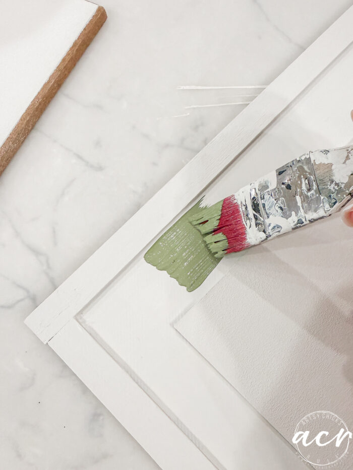 A hand uses a paintbrush to apply light green paint to the white wooden frame of a canvas, on a marble surface&mdash;perfect for your Easter Edition of Flipping Smalls projects.