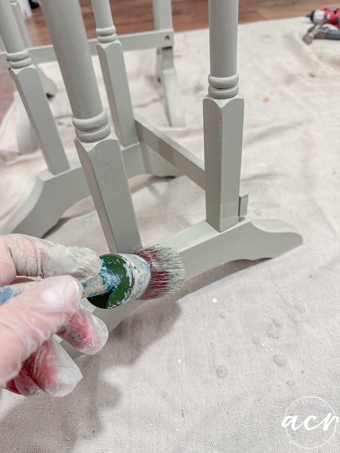 A gloved hand uses a round paintbrush to apply light green paint to the leg and support of a wooden table. Glass topped nesting tables and other painting supplies are visible on a protective drop cloth in the background.