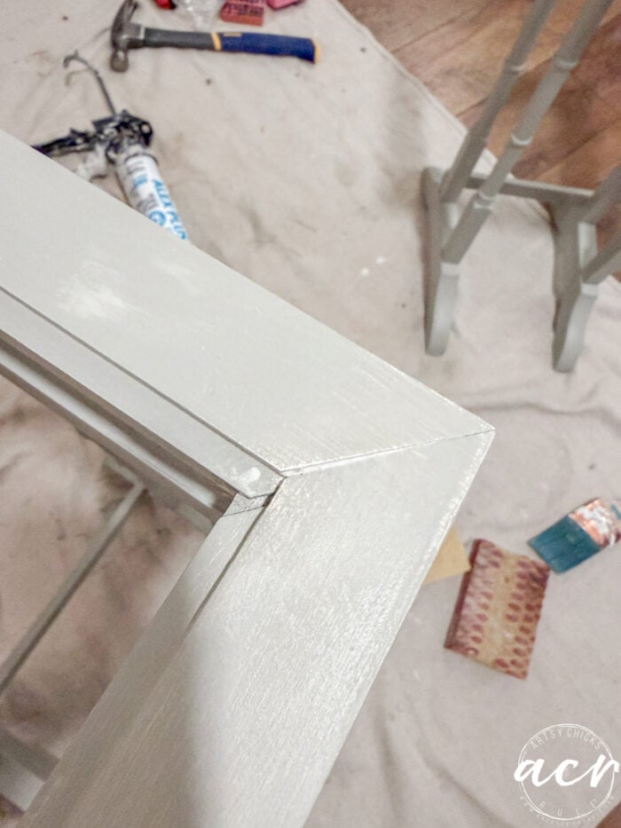 A close-up of a freshly painted, light-colored wooden frame corner, with painting and sanding supplies scattered on a drop cloth in the background&mdash;perfect inspiration for refurbishing glass top tables or stylish nesting tables.