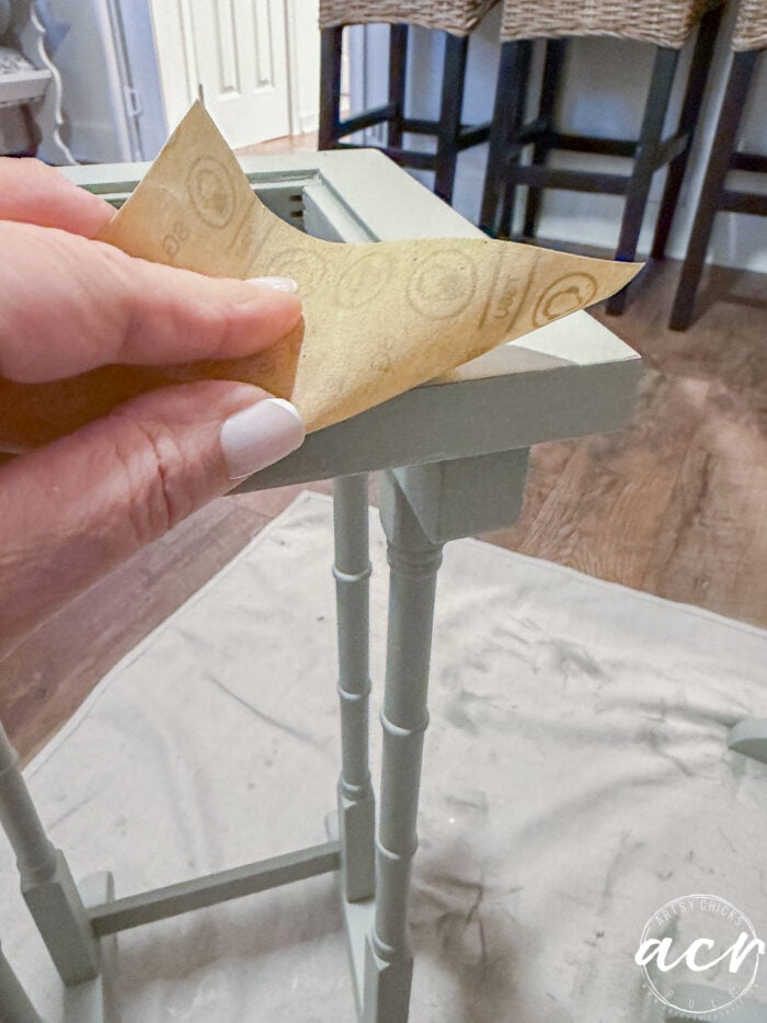 A hand with light-colored nail polish is holding sandpaper and sanding the edge of a small, light green nesting table set on a drop cloth indoors.