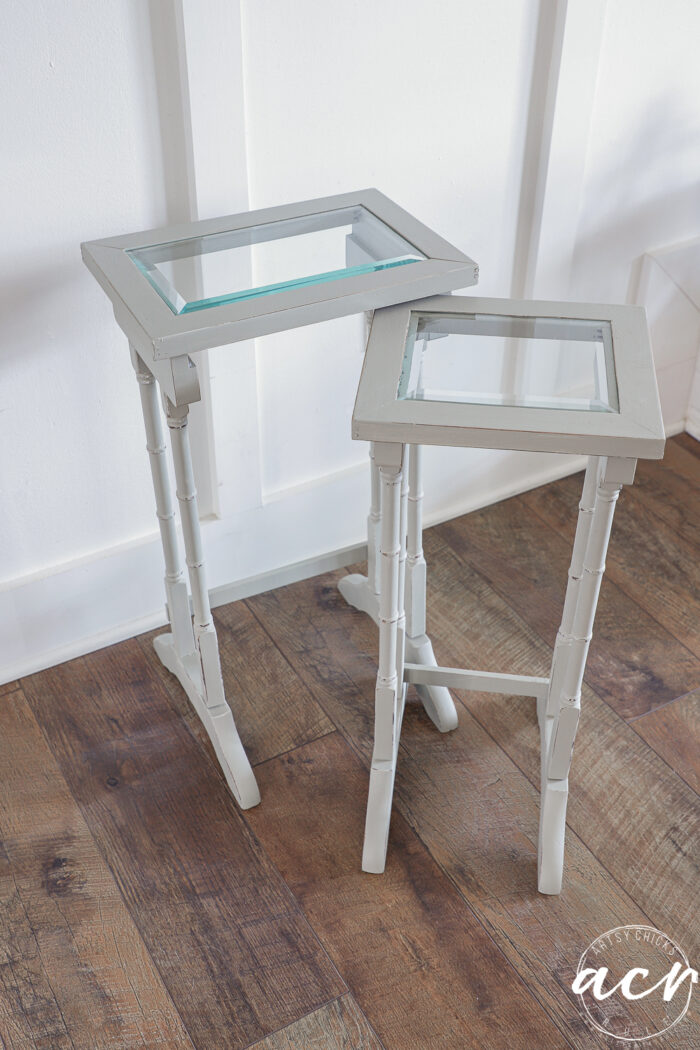 Two small, light gray nesting tables with glass tops and faux bamboo-style legs stand on a wooden floor against a white wall. The glass topped tables are different heights and overlap slightly for a chic table update.
