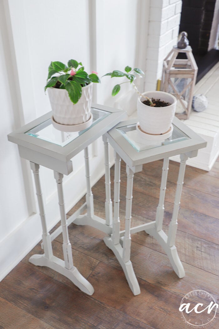 Two small, light gray glass topped nesting tables hold potted plants in white containers. The tables sit on a wood floor near a white wall, with a decorative lantern visible in the background.