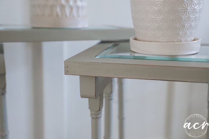 Close-up of two light green glass topped nesting tables with slightly distressed edges. Each table holds a white textured plant pot, while the softly blurred background enhances their charm.