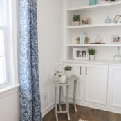 A cozy, bright corner features white built-in shelves with decor, a window with blue patterned curtains, glass topped nesting tables with potted plants, and a white and blue rug on wooden flooring.