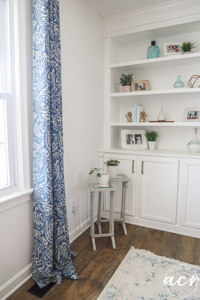 A cozy, bright corner features white built-in shelves with decor, a window with blue patterned curtains, glass topped nesting tables with potted plants, and a white and blue rug on wooden flooring.