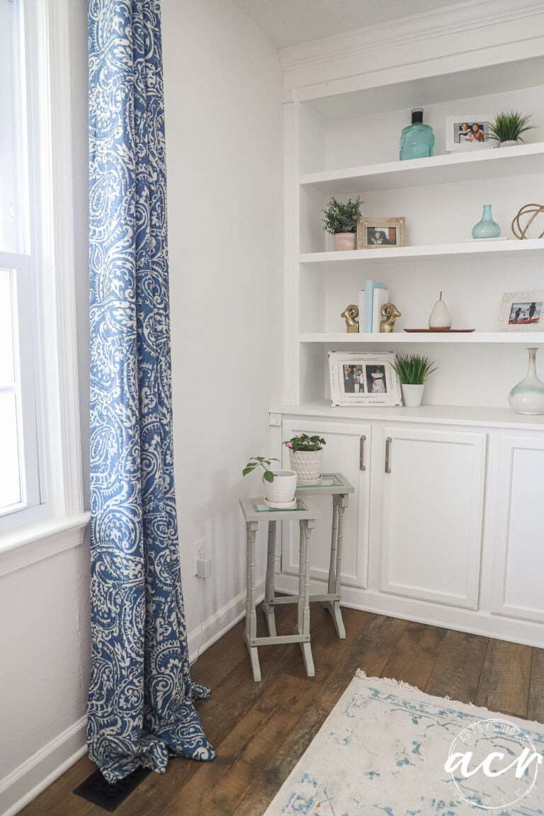 A cozy, bright corner features white built-in shelves with decor, a window with blue patterned curtains, glass topped nesting tables with potted plants, and a white and blue rug on wooden flooring.