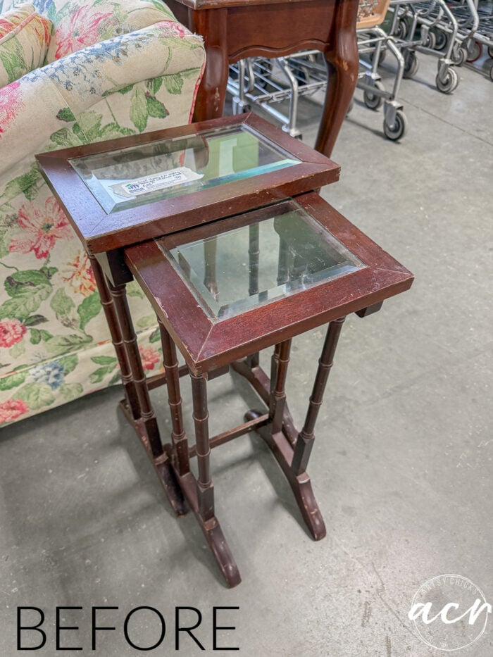 Two small Glass Topped Nesting Tables are stacked together in a thrift store, placed on a concrete floor near a floral upholstered chair and metal shopping carts. The word BEFORE appears in the corner.