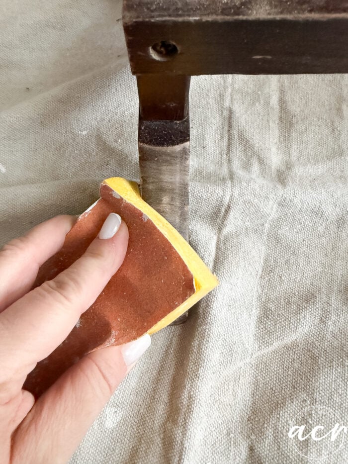 A hand holding sandpaper is smoothing the leg of a wooden piece of furniture, possibly part of glass topped nesting tables, which rests on a light-colored fabric surface.