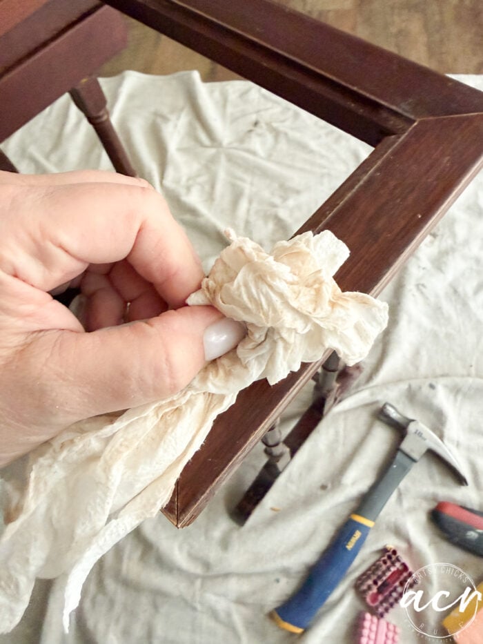 A hand uses a crumpled paper towel to clean a wooden nesting table leg; tools including a hammer and pliers are visible on a cloth-covered floor in the background.