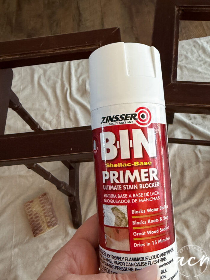 A hand holds a can of Zinsser B-I-N Shellac-Base Primer in front of a wooden frame, with a sanding sponge and brown paper on a light surface&mdash;perfect for refinishing your glass topped nesting tables.