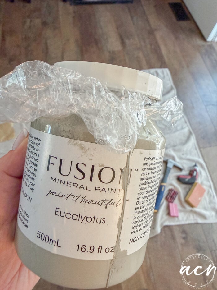 A hand holds a 500mL jar of Fusion Mineral Paint in Eucalyptus, with plastic wrap under the lid. Paint supplies and a drop cloth surround Glass Topped Nesting Tables on a wooden floor in the background.