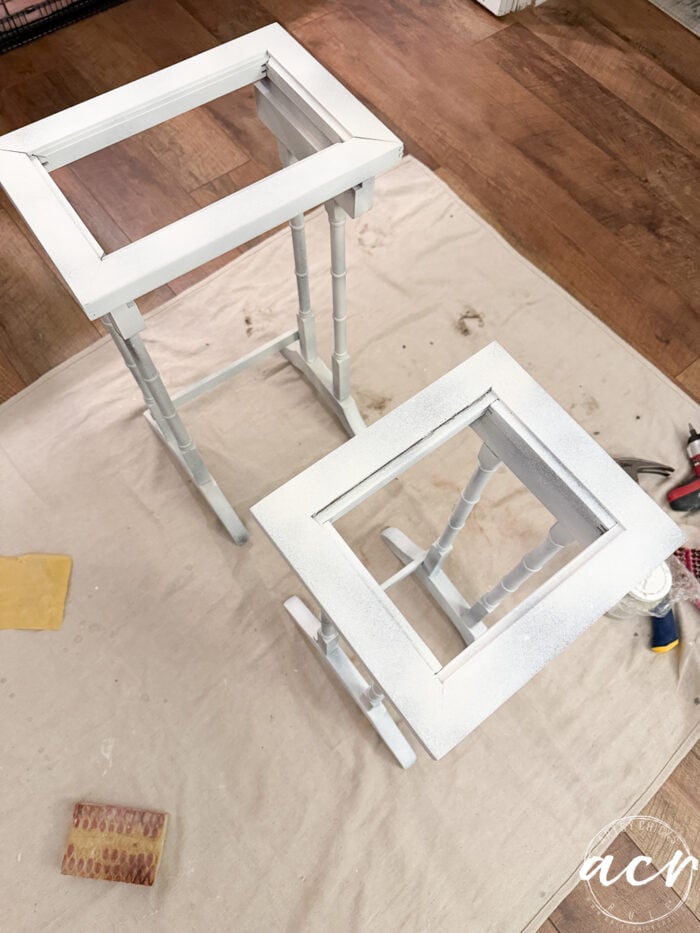 Two small wooden nesting tables with square tops are being painted white, positioned on a drop cloth with sanding blocks, pliers, and paint supplies nearby, on a wood floor.