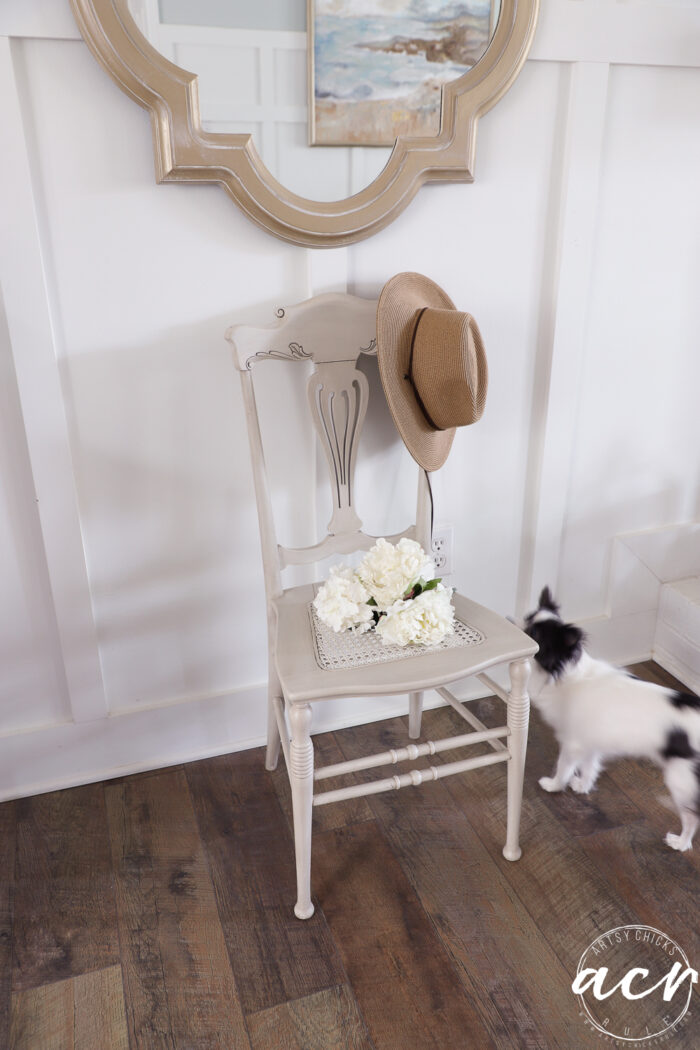 A beige Vintage Chair with a straw hat on the backrest and a tray of white flowers on the seat sits against a white paneled wall; nearby, a small black and white dog stands on the wooden floor&mdash;a charming scene for any Vintage Chair Update.