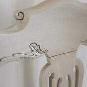 Close-up of the top section of a light-colored, ornately carved vintage chair with decorative scroll and leaf patterns. A round watermark with the initials acn appears in the lower right corner.