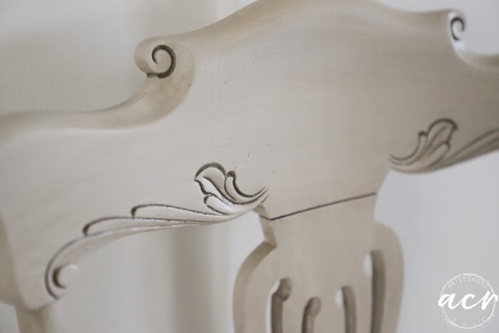 Close-up of the top section of a light-colored, ornately carved vintage chair with decorative scroll and leaf patterns. A round watermark with the initials acn appears in the lower right corner.