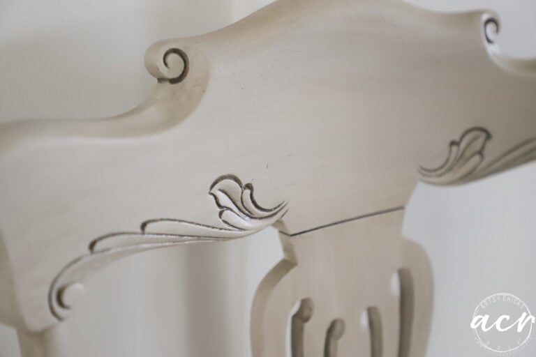 Close-up of the top section of a light-colored, ornately carved vintage chair with decorative scroll and leaf patterns. A round watermark with the initials acn appears in the lower right corner.