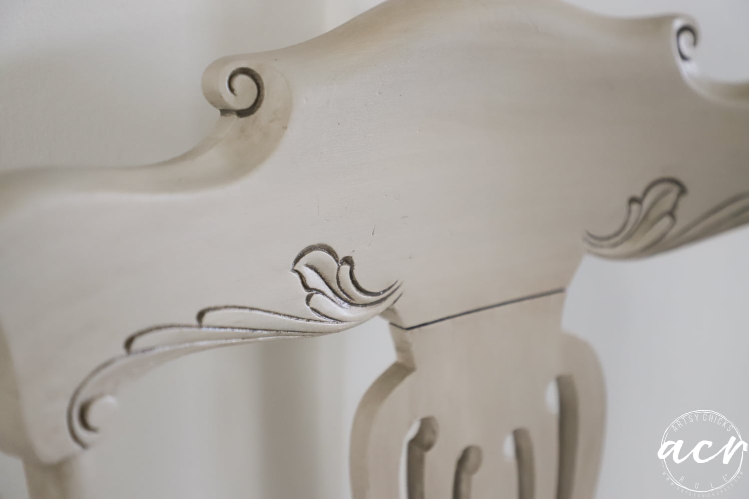 Close-up of the top section of a light-colored, ornately carved vintage chair with decorative scroll and leaf patterns. A round watermark with the initials acn appears in the lower right corner.