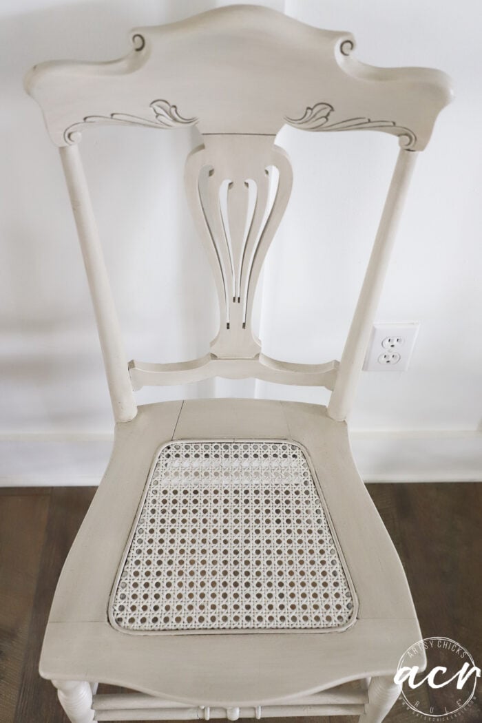 A vintage chair with ornate carvings on the backrest and a cane seat sits against a white wall and dark wood floor. An electrical outlet is visible on the wall behind this timeless piece.