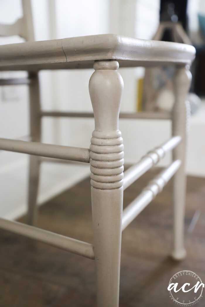 Close-up of a light-colored, painted wooden chair leg with carved details and a smooth finish, showcasing vintage chair restoration on a wooden floor, with part of another vintage chair visible in the background.