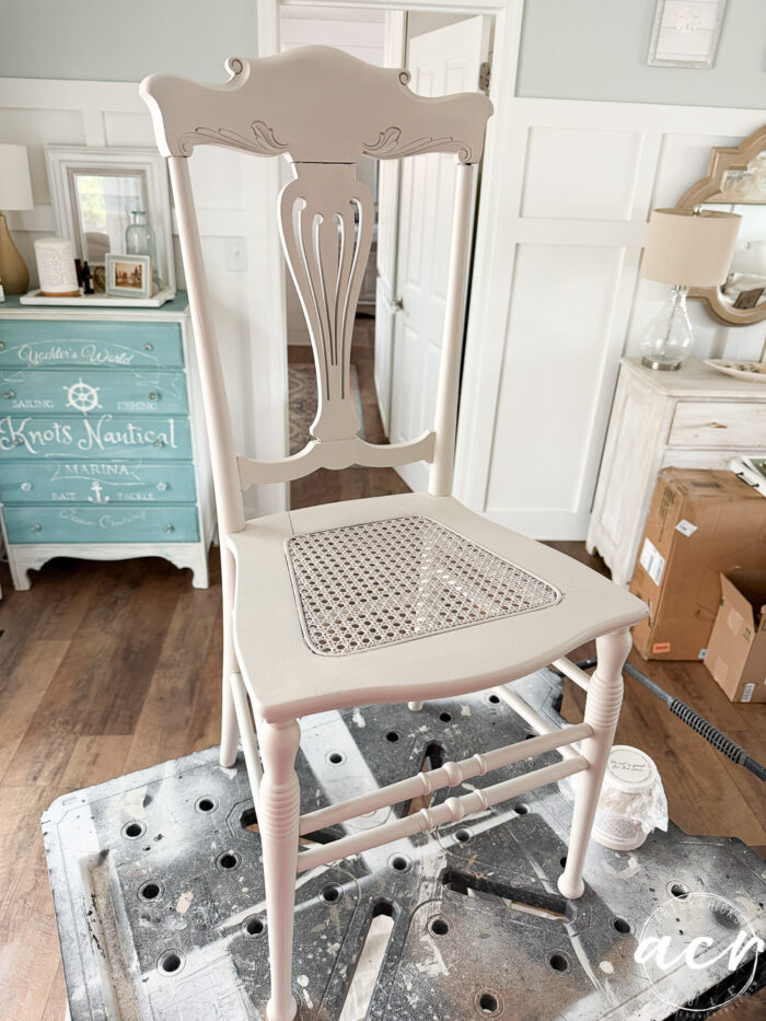 A vintage chair with a pale beige paint finish and a cane seat sits on a workbench in a bright, airy room with cottage-style decor&mdash;the perfect setting for your next charming vintage furniture update.