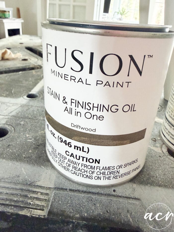 A can of Fusion Mineral Paint Stain & Finishing Oil (Driftwood) sits on a work surface, ready for your next vintage chair update, showing the label with product details and safety cautions.