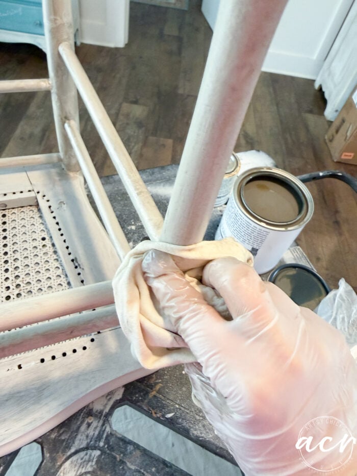 A gloved hand uses a cloth to apply finish to the leg of an upside-down vintage chair; an open can of paint or varnish and supplies are visible nearby, highlighting a careful chair update in progress.