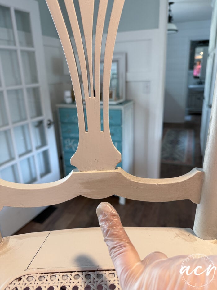 A gloved finger points at a detail on the backrest of a partially painted vintage chair, with a light blue cabinet and glass-paneled door visible in the background&mdash;a charming chair update in progress.