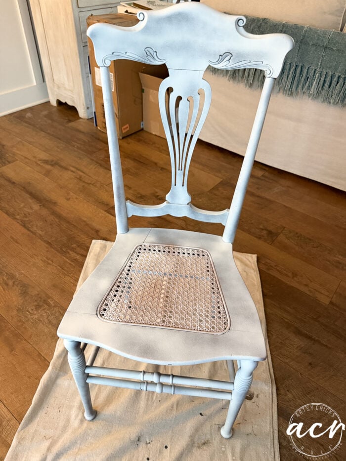 A vintage chair with a cane seat is partially painted in light blue and white, sitting on a drop cloth on a hardwood floor, with a sofa and blanket in the background&mdash;perfect for your next chair update.