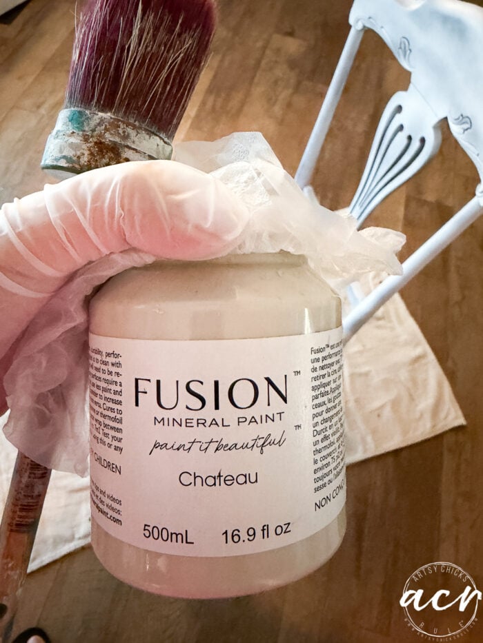 A gloved hand holds a jar of Fusion Mineral Paint in Chateau with a paintbrush, beside a partially painted blue and white vintage chair on a wooden floor&mdash;perfect for your next vintage furniture update.