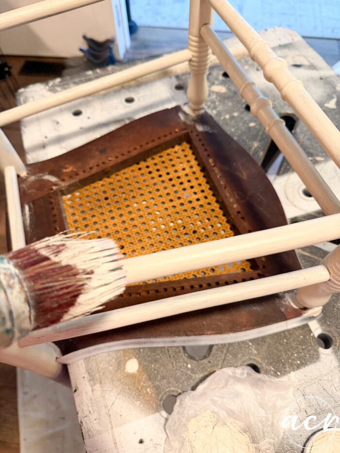 A close-up of a hand painting the wooden frame of an upside-down vintage chair white. The chair has a woven cane seat, and paint supplies are visible on the work surface underneath, showcasing a charming chair update in progress.