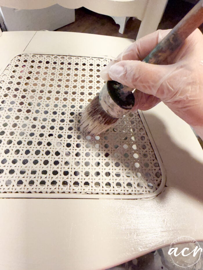 A gloved hand uses a round paintbrush to update a vintage chair seat with a light cream color. The intricate woven pattern of this vintage furniture piece is clearly visible.