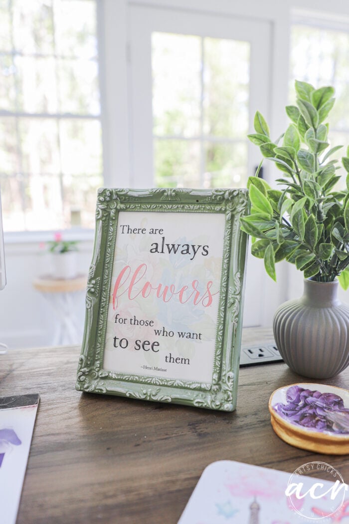 A decorative picture frame with a quote sits on a wooden desk in this sunroom-inspired office reveal, next to a small potted plant. The quote reads, There are always flowers for those who want to see them. Sunlight streams through the window behind.