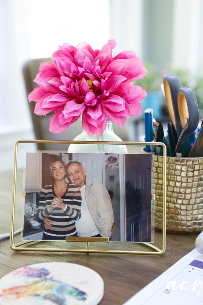 A framed photo of two smiling people sits on a desk next to a vase with a large pink flower and a gold holder with pens and scissors. The cozy workspace, perfect for an office reveal, feels both welcoming and inspiring.