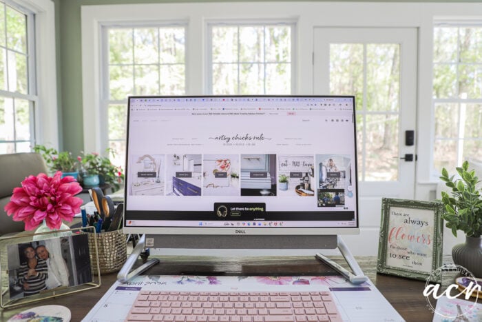 A computer monitor on a desk displays a home decor blog. The sunroom desk features pink flowers, framed photos, a calendar, and decor items. Large windows in the background fill this inspiring Office Reveal with natural light.