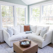 A bright sunroom with large windows, a white sectional sofa adorned with throw pillows, a tan ottoman topped by a wicker tray holding magazines, and a patterned rug on the floor&mdash;perfect for your next Office Reveal.