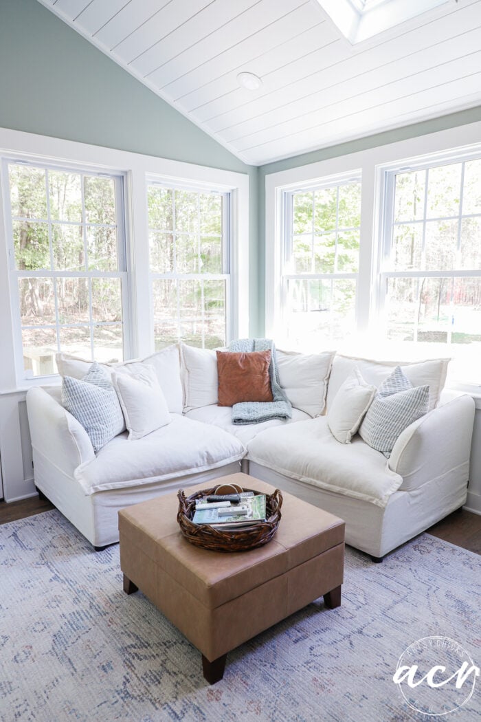 A bright sunroom with large windows, a white sectional sofa adorned with throw pillows, a tan ottoman topped by a wicker tray holding magazines, and a patterned rug on the floor&mdash;perfect for your next Office Reveal.