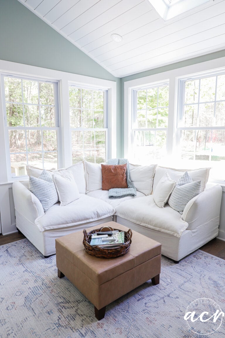 A bright sunroom with large windows, a white sectional sofa adorned with throw pillows, a tan ottoman topped by a wicker tray holding magazines, and a patterned rug on the floor&mdash;perfect for your next Office Reveal.
