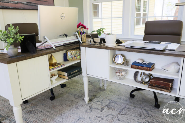 Two white desks arranged in an L-shape in a bright sunroom office, each with a chair, computer, plants, books, and decorative items. Windows flood the space with natural light, while a patterned rug completes this stylish office reveal.