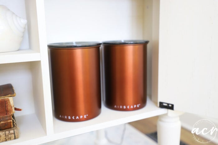 Two metallic brown canisters labeled Airscrape sit side by side on a white shelf in the sunroom, with books and a shell visible nearby.