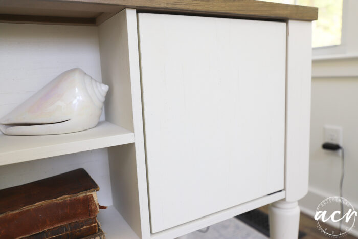 A white wooden cabinet with a closed door sits in a sunroom, a shiny conch shell on its shelf and a couple of old brown books below. Light from a window brightens the scene.