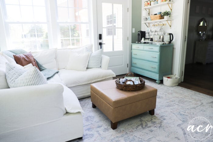 Bright living room doubles as a sunroom, with a white sofa, beige ottoman with wicker tray, blue sideboard with coffee supplies, open shelves with cups, and large windows. Cozy, airy, and inviting decor perfect for an office reveal.